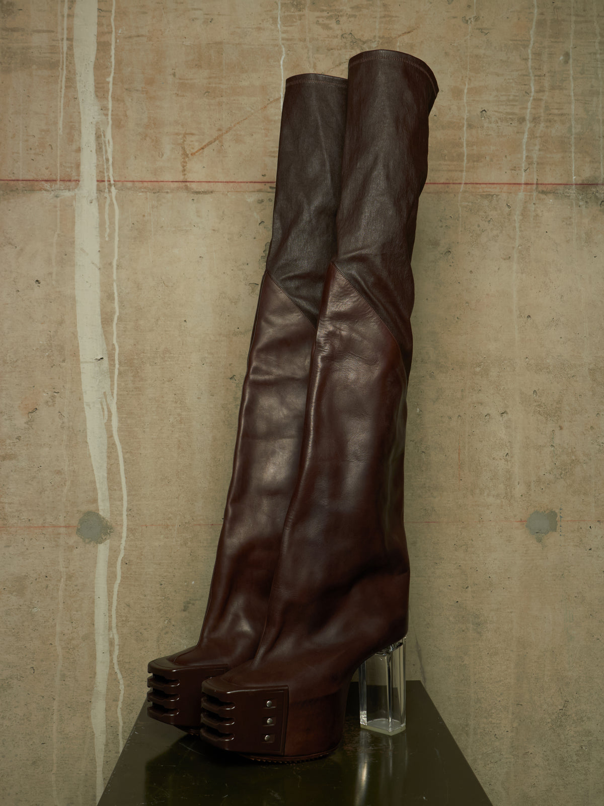 A pair of dark brown leather over-the-knee boots with chunky platform soles and clear acrylic block heels, displayed on a dark surface against a textured concrete wall.