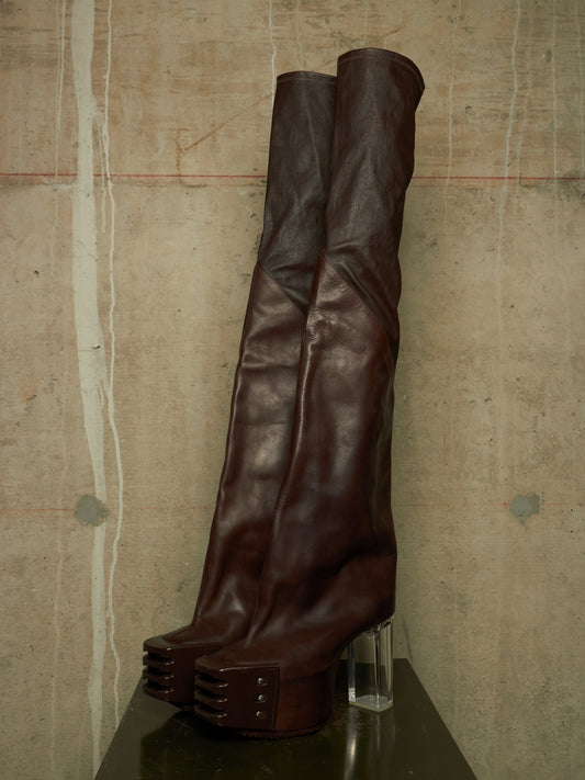 A pair of dark brown leather over-the-knee boots with chunky platform soles and clear acrylic block heels, displayed on a dark surface against a textured concrete wall.