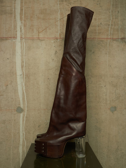 A single dark brown leather over-the-knee boot with a chunky platform sole and clear acrylic block heel, displayed on a dark reflective surface against a textured concrete wall.
