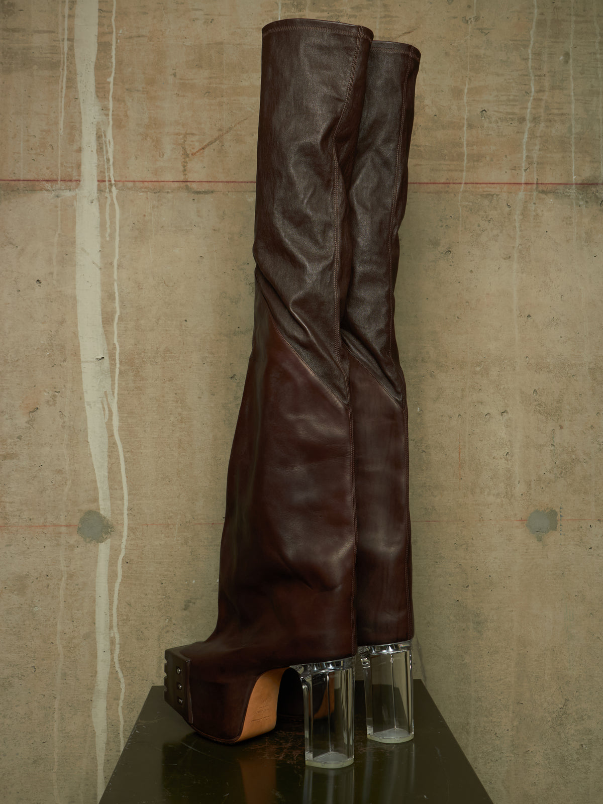 A pair of dark brown leather over-the-knee boots with chunky platform soles and clear acrylic block heels, displayed on a dark reflective surface against a textured concrete wall.