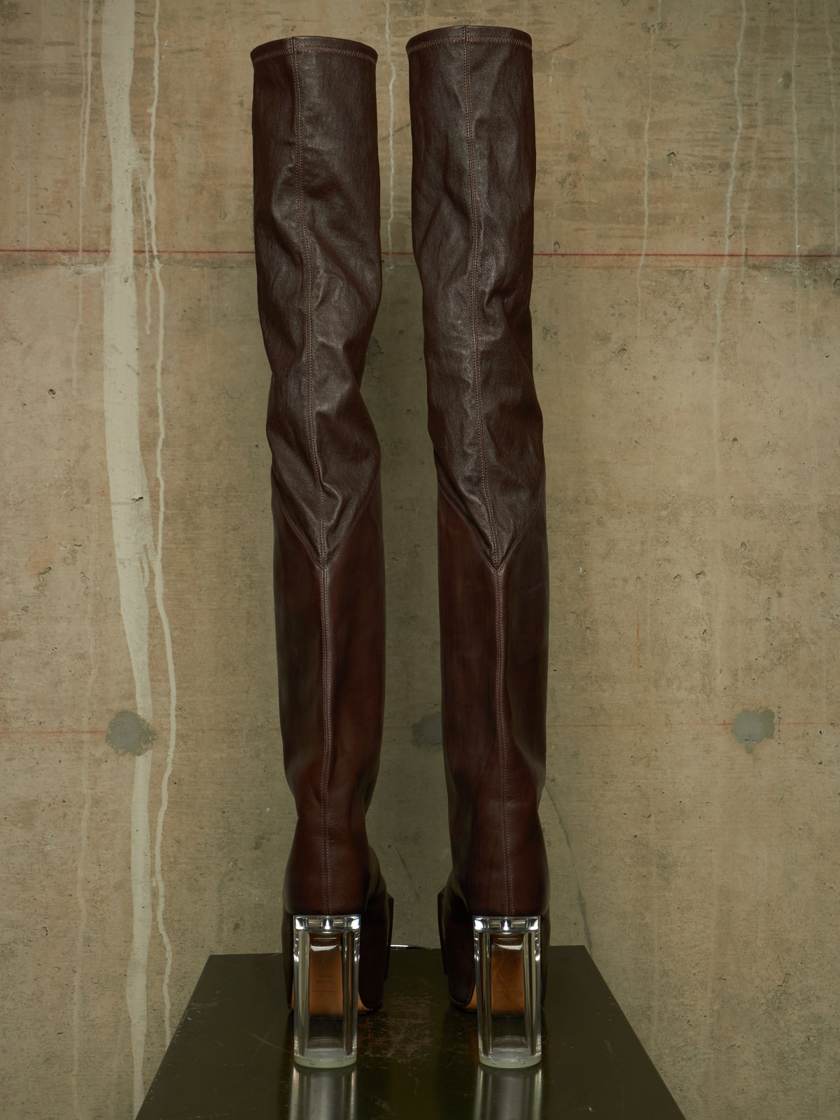 A pair of dark brown leather over-the-knee boots with clear acrylic block heels, standing on a dark reflective surface against a textured concrete wall.