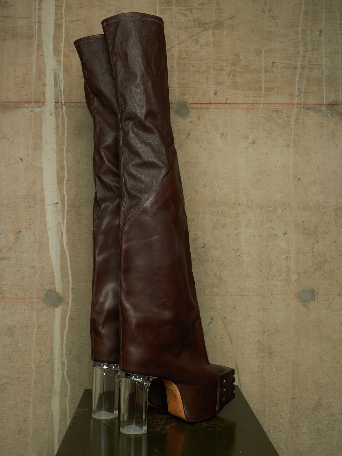 A pair of dark brown leather over-the-knee boots with clear cylindrical block heels, displayed on a dark reflective surface against a textured concrete wall.