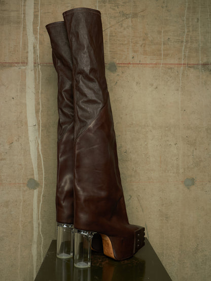 A pair of dark brown leather over-the-knee boots with clear cylindrical block heels, displayed on a dark reflective surface against a textured concrete wall.