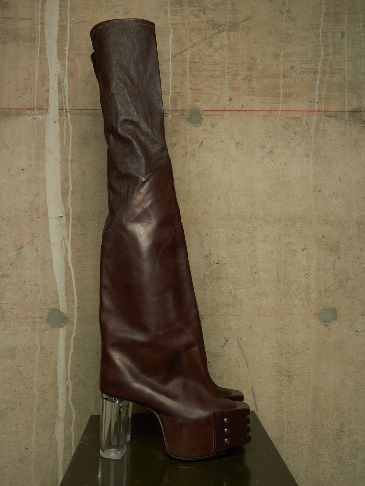A dark brown leather over-the-knee boot with a chunky clear acrylic block heel and a platform sole with metal studs, displayed on a dark reflective surface against a textured concrete wall.