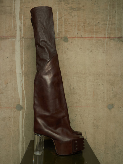 A dark brown leather over-the-knee boot with a chunky clear acrylic block heel and a platform sole with metal studs, displayed on a dark reflective surface against a textured concrete wall.