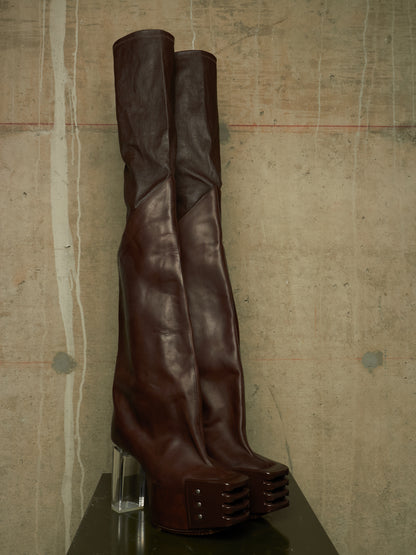 A pair of dark brown leather over-the-knee boots with a chunky clear acrylic block heel and stacked platform sole, displayed on a dark surface against a textured concrete wall.