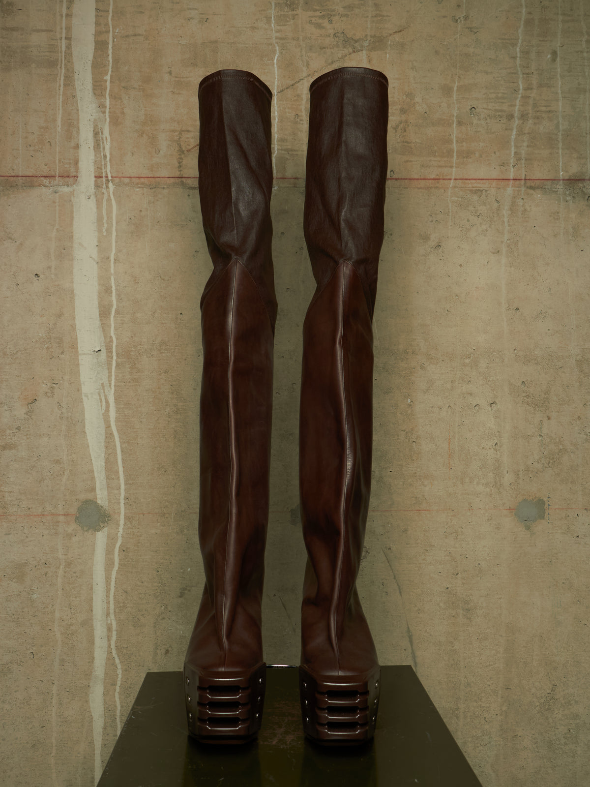 A pair of dark brown leather over-the-knee boots with sculptural, twisted shafts and chunky platform heels, displayed on a black pedestal against a textured concrete wall.