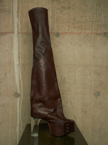 A dark brown leather over-the-knee boot with a chunky clear acrylic block heel and a platform sole with metal studs, displayed on a dark surface against a textured concrete wall.