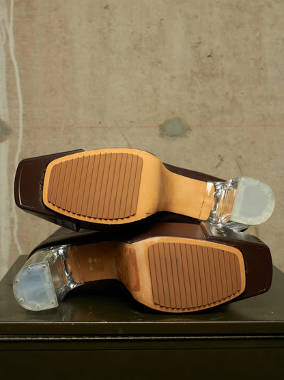 A pair of brown leather shoes shown from the sole, featuring tan rubberized tread patterns and clear plastic protective caps on the heels, resting on a dark surface against a textured beige wall.