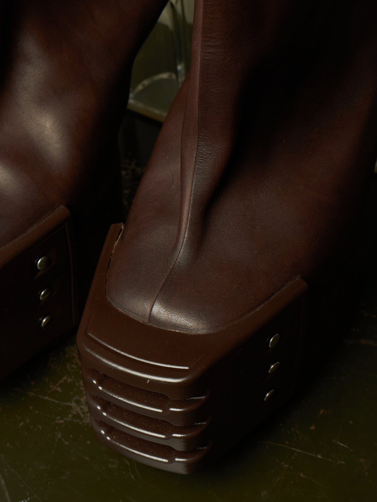 Close-up of dark brown leather platform boots with chunky lug soles and metal rivet details, photographed in dim lighting on a dark floor.