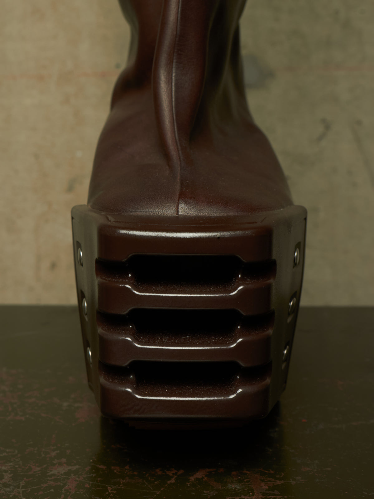 A close-up rear view of a dark brown leather boot with an extremely chunky, sculptural platform heel featuring horizontal ridges and side rivets, set against a textured neutral background.