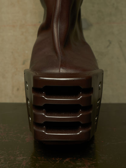 A close-up rear view of a dark brown leather boot with an extremely chunky, sculptural platform heel featuring horizontal ridges and side rivets, set against a textured neutral background.