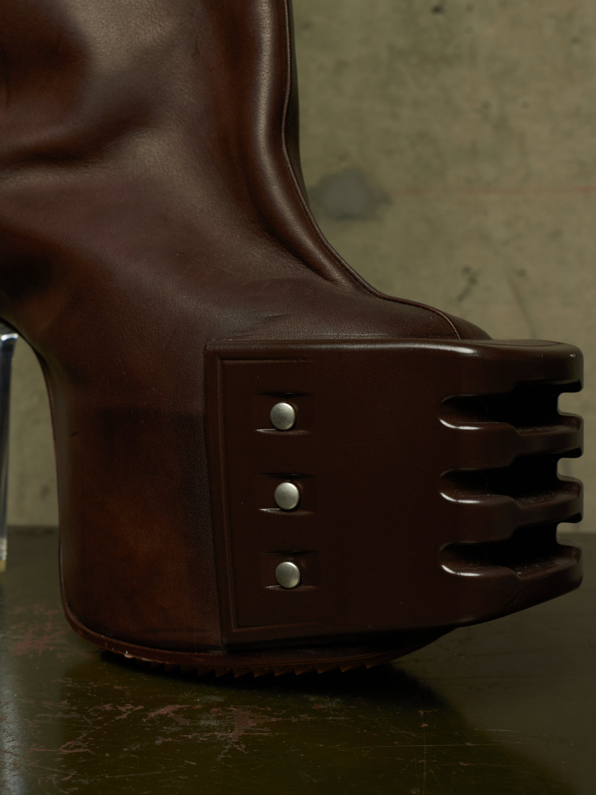 A close-up of a dark brown leather boot with a highly sculptural, ribbed platform sole and three silver rivets on the side.
