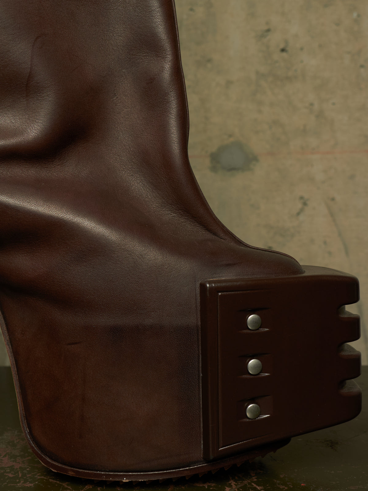 Close-up of a dark brown leather boot with a chunky sculptural platform heel and metal rivet details, set against a textured concrete background.