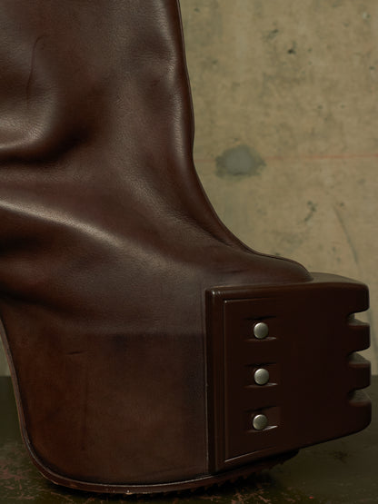 Close-up of a dark brown leather boot with a chunky sculptural platform heel and metal rivet details, set against a textured concrete background.