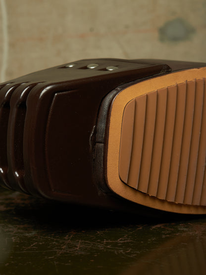 Close-up detail of a dark brown leather shoe sole showing the ribbed tan outsole and small metal eyelets, resting on a dark surface.