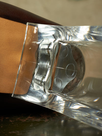 A close-up of a clear acrylic or glass tumbler with a built-in metal cup holder and silver cup, resting against a brown leather surface.