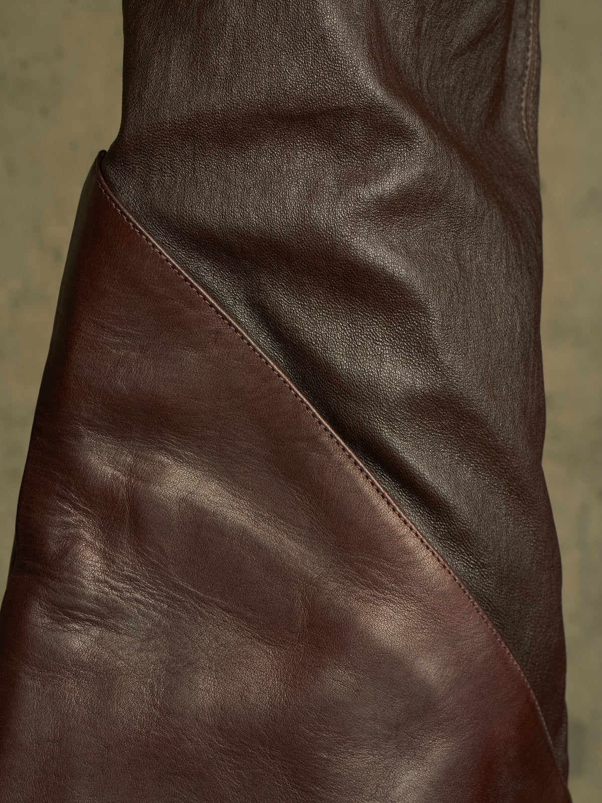 Close-up of a dark brown leather garment, showing the texture and diagonal seam against a neutral background.