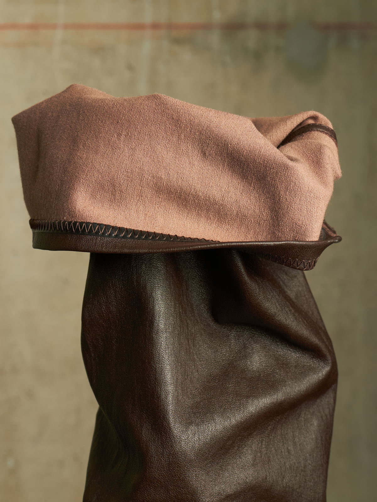 A close-up of a brown leather boot or shoe with a folded tan suede or wool cuff, set against a textured neutral background.
