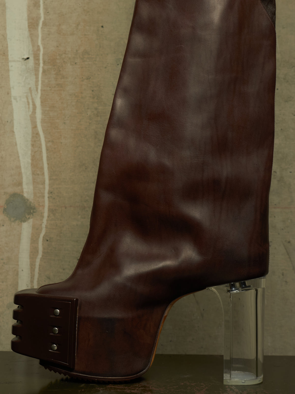 A dark brown leather platform boot with a clear cylindrical block heel, shown against a textured concrete wall background.