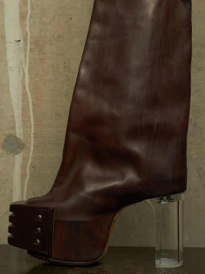 A dark brown leather platform boot with a clear cylindrical block heel, shown against a textured concrete wall background.
