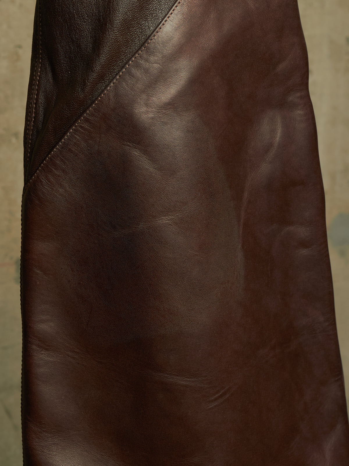 A close-up of a dark brown leather garment, showing the smooth texture and diagonal seam stitching.