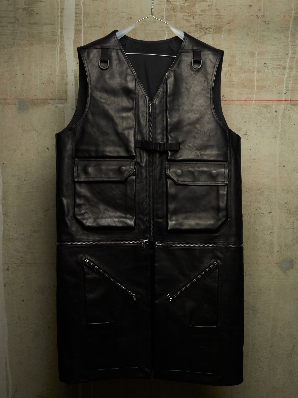 A black leather utility vest with multiple zippered and flap pockets, hanging on a clear plastic hanger against a textured concrete wall.