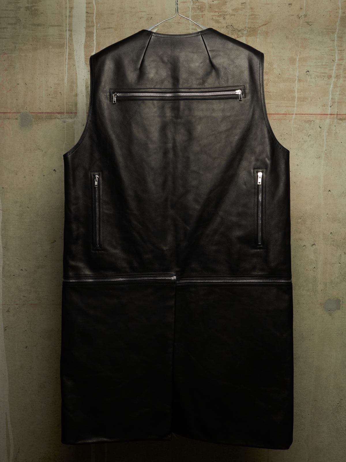 A black leather sleeveless vest with silver zippers, hanging on a wire hanger against a textured concrete wall.
