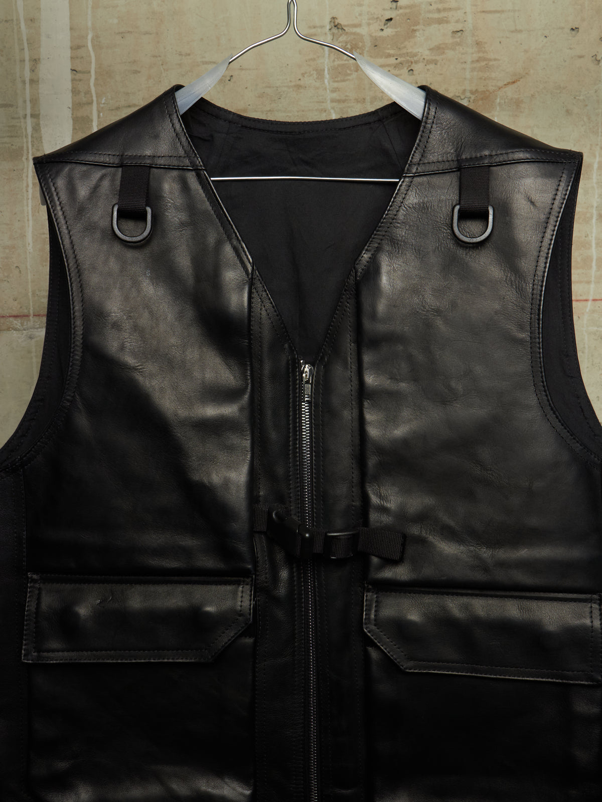 Black leather motorcycle vest with a front zipper, side D-ring straps, and flap pockets, hanging on a metal hanger against a textured beige wall.