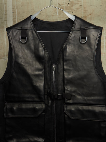 Black leather motorcycle vest with a front zipper, side D-ring straps, and flap pockets, hanging on a metal hanger against a textured beige wall.