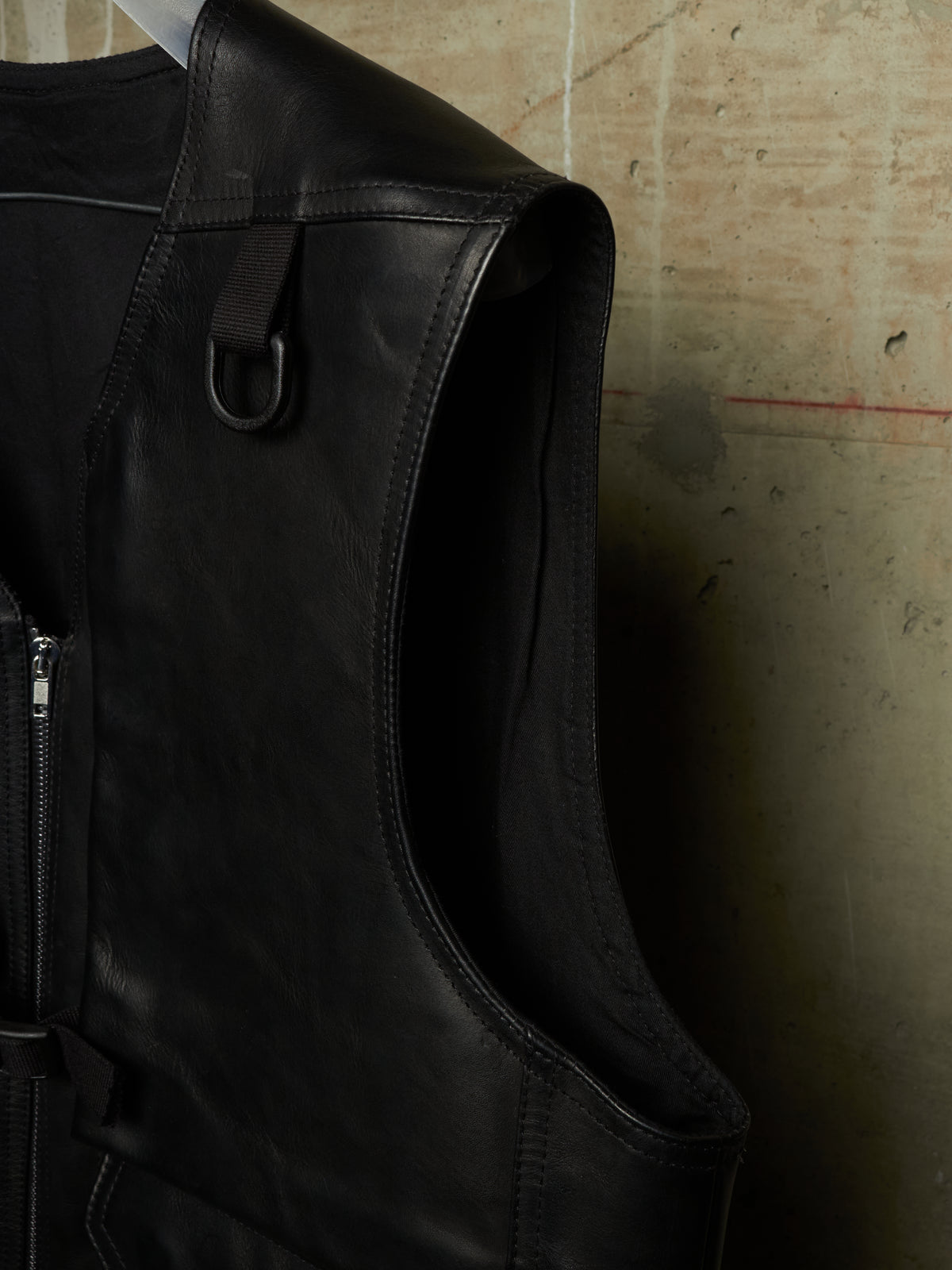 Close-up of a black leather vest with a side zipper and a hanging D-ring, set against a textured concrete wall.
