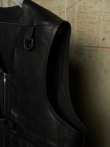 Close-up of a black leather vest with a side zipper and a hanging D-ring, set against a textured concrete wall.