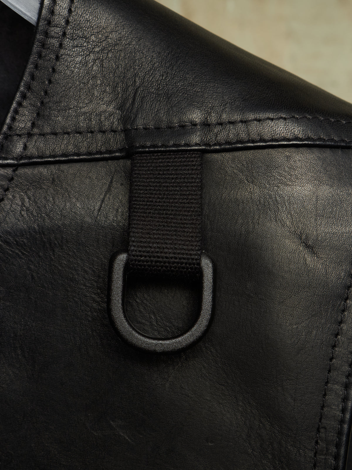 Close-up detail of a black leather bag showing a black webbing strap attached to a black metal D-ring, with visible stitching and leather texture.