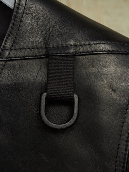 Close-up detail of a black leather bag showing a black webbing strap attached to a black metal D-ring, with visible stitching and leather texture.