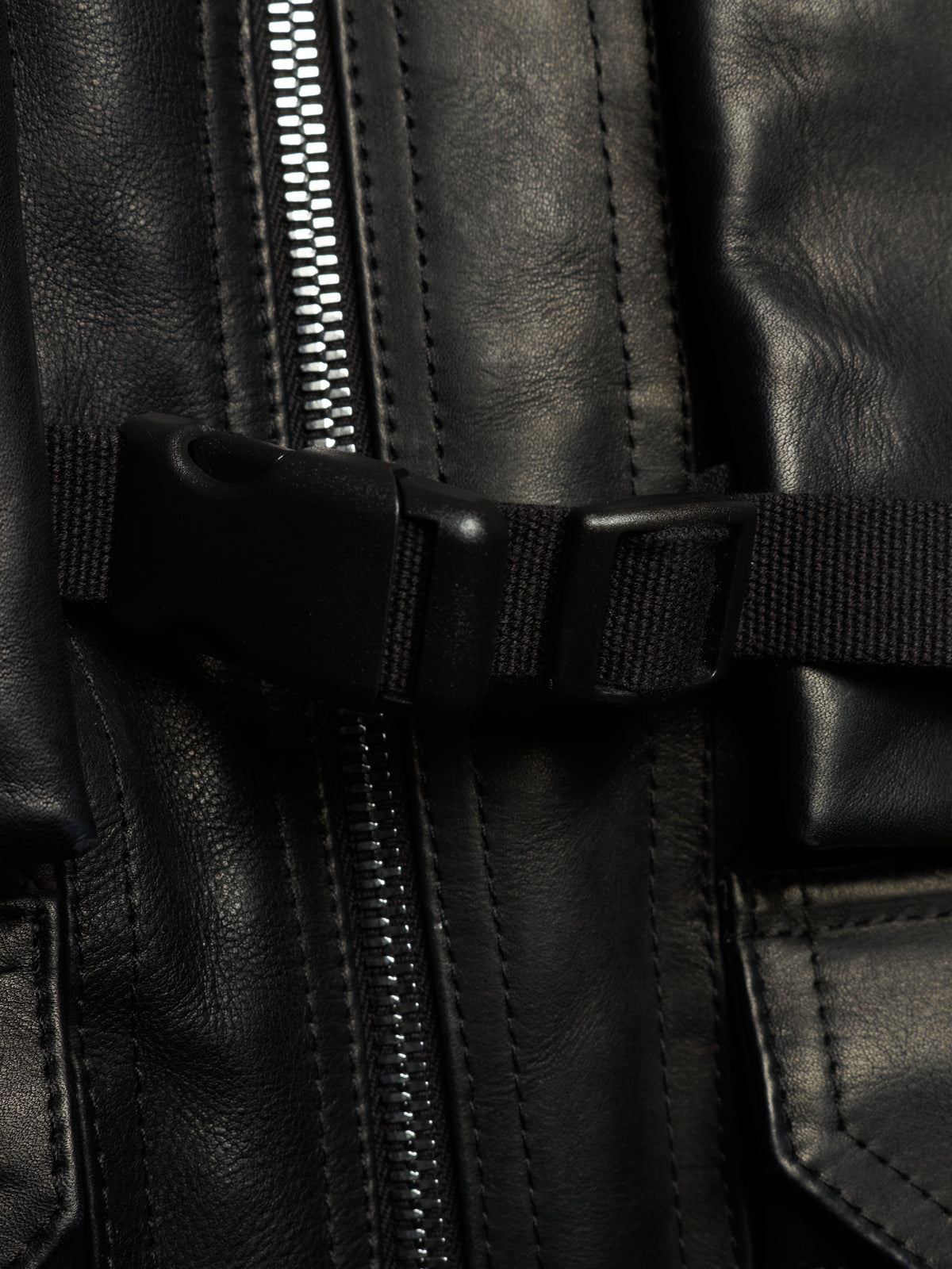 Close-up detail of a black leather jacket featuring a central silver zipper, a black webbing strap with a plastic buckle, and stitched leather panels.