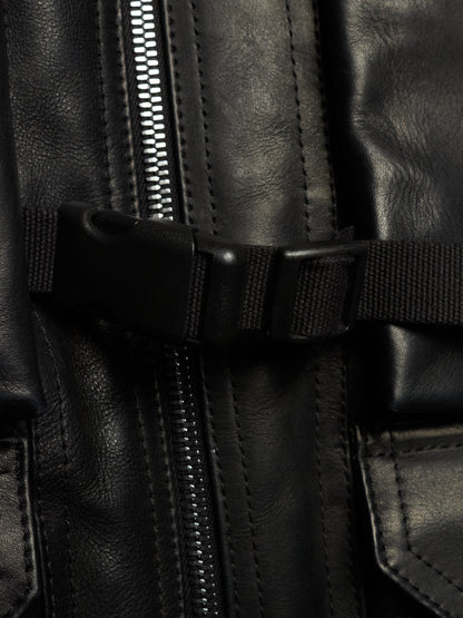Close-up detail of a black leather jacket featuring a central silver zipper, a black webbing strap with a plastic buckle, and stitched leather panels.