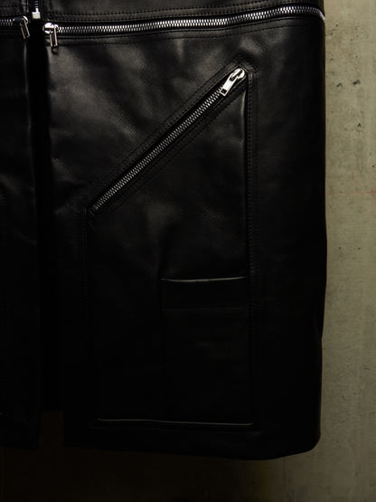 Close-up of a black leather jacket or bag with diagonal zippered pockets and silver zipper hardware, photographed against a textured gray wall.