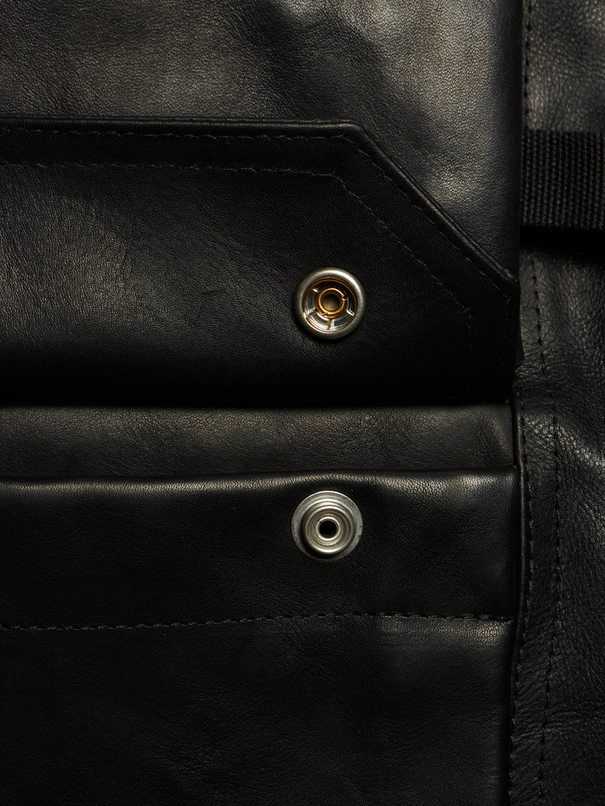 Close-up of a black leather product showing two silver metal snap buttons and fine stitching details.