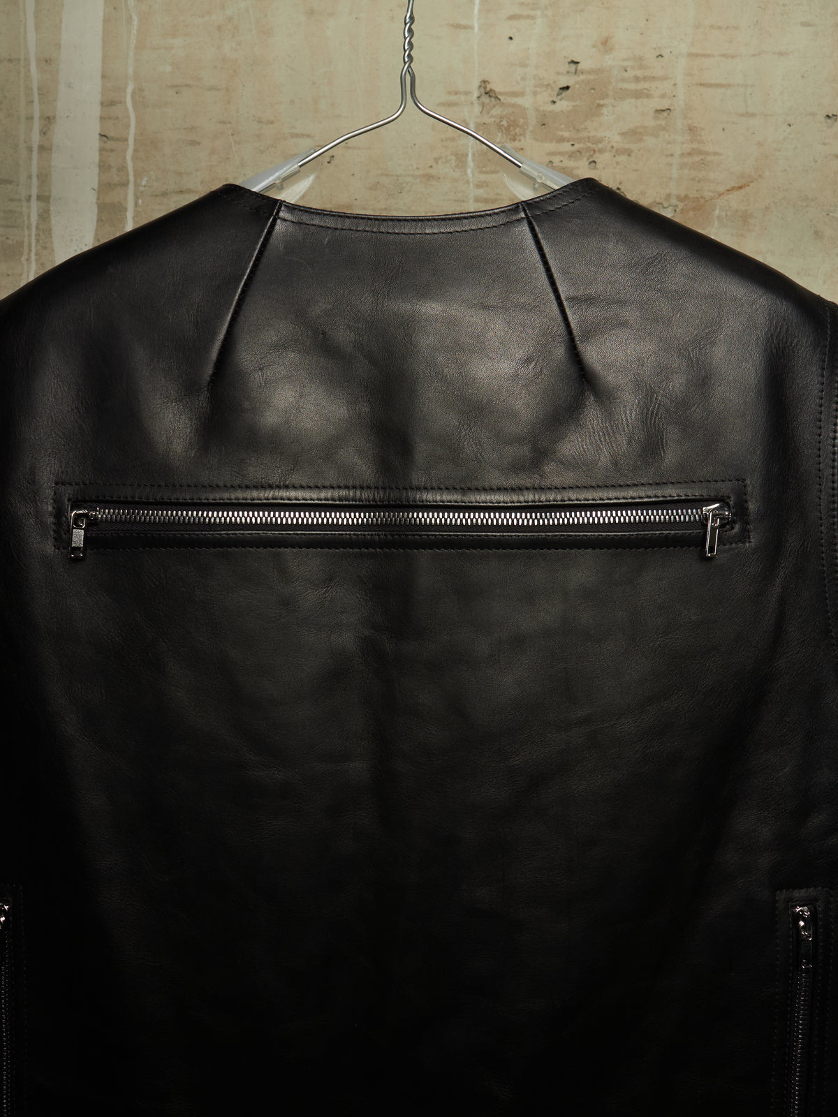 A black leather jacket hanging on a metal hanger against a textured beige wall, showing a horizontal zippered pocket on the back.