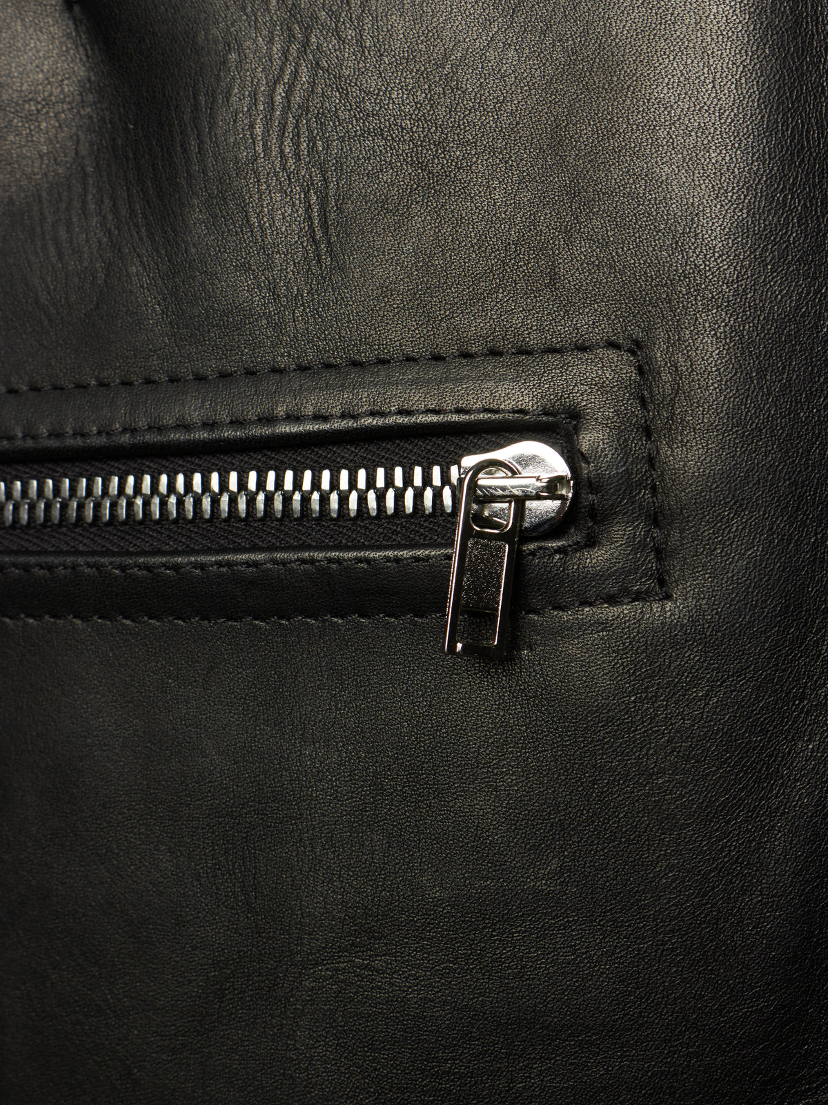 Close-up of a black leather backpack or bag featuring a silver metal zipper with a pull tab, set against a dark textured leather background.
