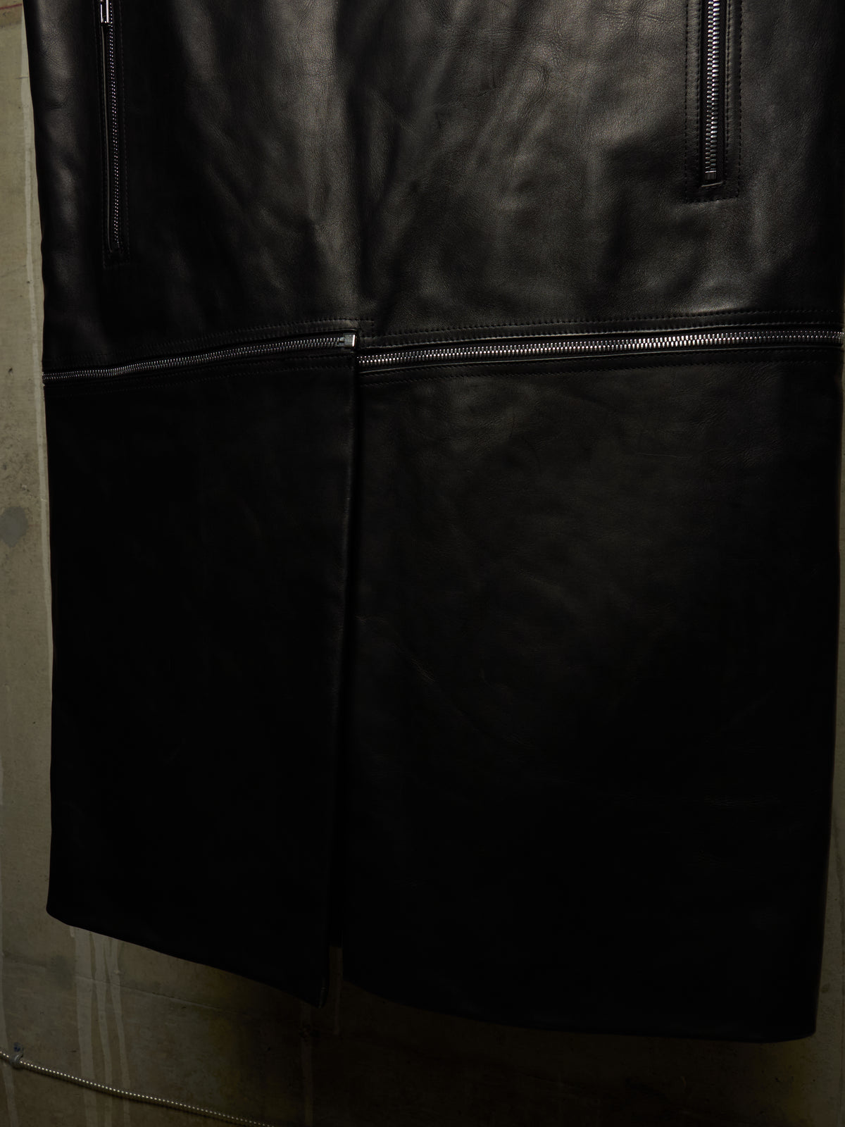Close-up of a black leather garment, likely a jacket or coat, showing the smooth texture, a central seam, and zipper details against a dark background.