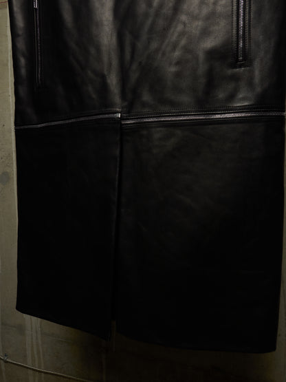 Close-up of a black leather garment, likely a jacket or coat, showing the smooth texture, a central seam, and zipper details against a dark background.