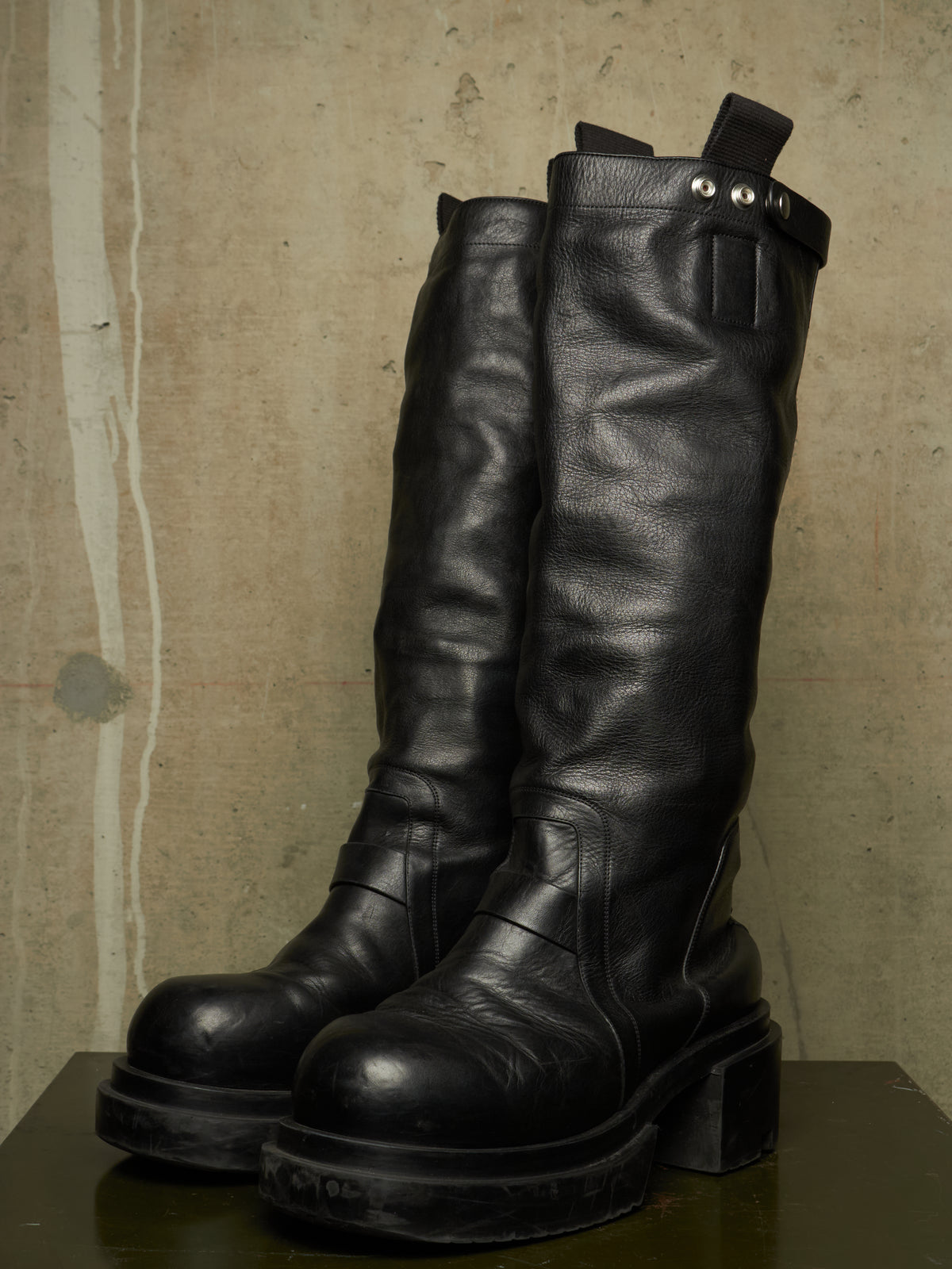 A pair of tall black leather boots with chunky block heels and silver rivets, displayed on a dark surface against a textured concrete wall.