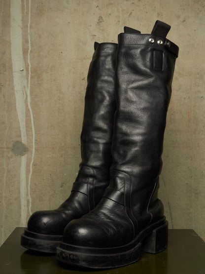 A pair of tall black leather boots with chunky block heels and silver rivets, displayed on a dark surface against a textured concrete wall.