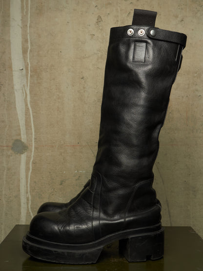 A single tall black leather boot with a chunky lug sole and block heel, photographed against a textured beige wall on a dark surface.