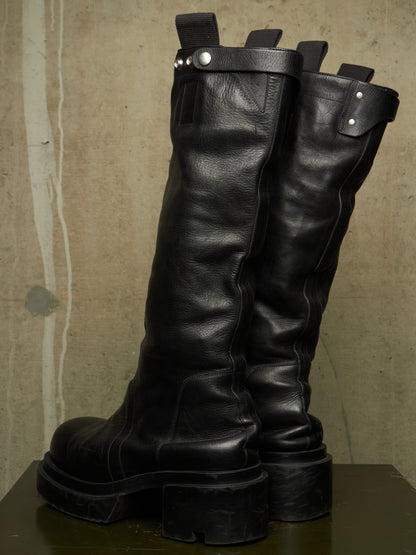 A pair of tall black leather boots with chunky lug soles and silver-tone hardware, displayed on a dark surface against a textured beige wall.