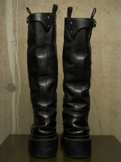 A pair of tall black leather boots with chunky platform soles and metal pull loops, standing upright on a dark surface against a textured beige wall.