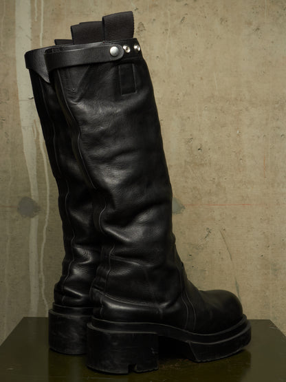 A pair of black leather knee-high boots with chunky lug soles and a decorative strap with silver studs, displayed on a dark surface against a textured concrete wall.