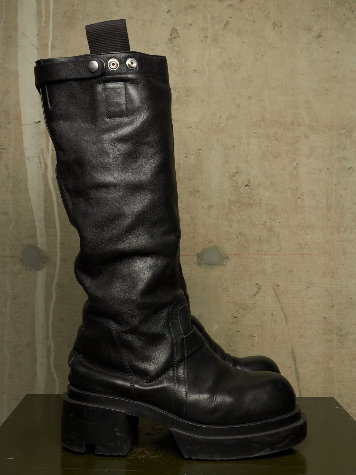 A single black leather knee-high boot with a chunky lug sole and metal snap details near the top, photographed against a textured concrete wall.