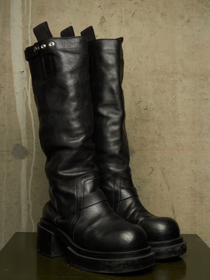 A pair of tall black leather boots with chunky platform soles and a buckle detail on the side, displayed on a dark surface against a textured concrete wall.